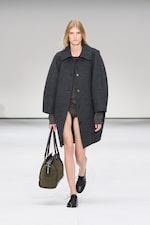 Placeholder for: Model in oversized dark coat, carrying a large olive bag, walks down a minimalist runway. Black shoes and neutral expression.
