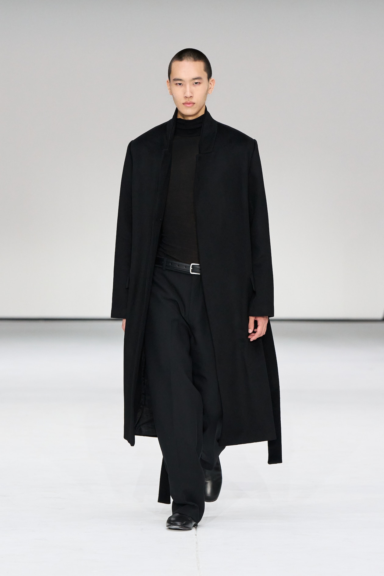 A model in a long black coat, black turtleneck, and pants walks confidently on a minimalist runway.