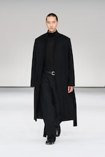 Placeholder for: A model in a long black coat, black turtleneck, and pants walks confidently on a minimalist runway.