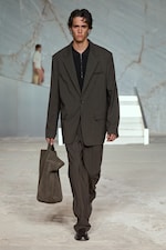 Placeholder for: Model in an oversized dark suit walks on a marble-patterned runway, carrying a large matching bag, with a distant figure in the background.