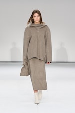 Placeholder for: Model in a plaid outfit with a long skirt and oversized top, carrying a matching bag, walking on a white runway.