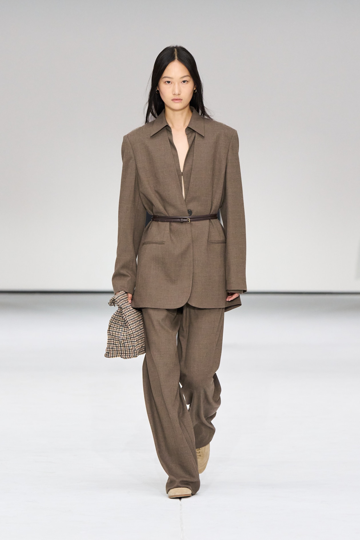 Model walking on runway in a brown oversized suit with a belt, holding a checkered bag. Minimalist background.