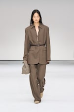 Placeholder for: Model walking on runway in a brown oversized suit with a belt, holding a checkered bag. Minimalist background.