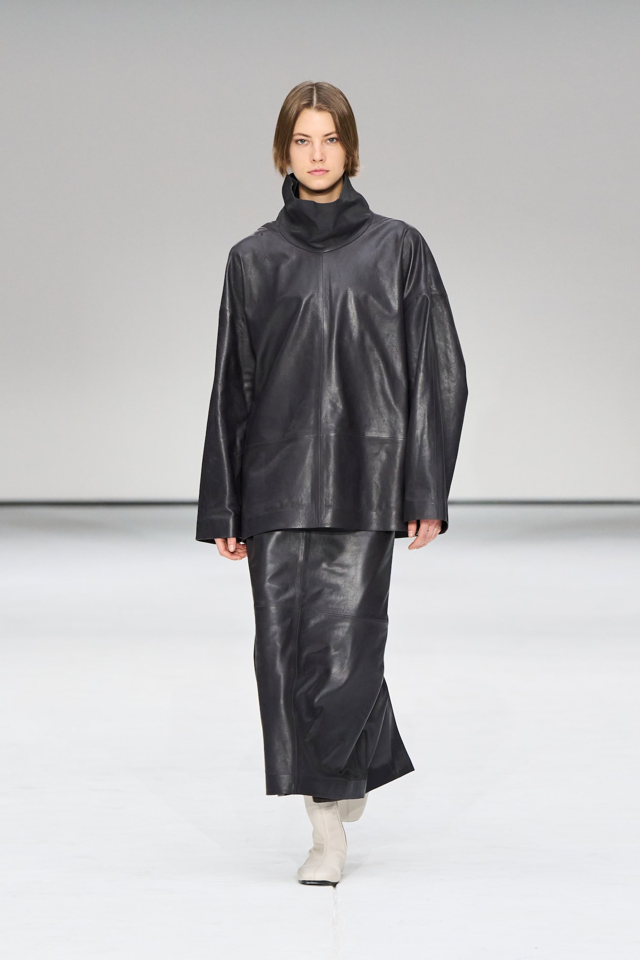Model wearing a black leather outfit with a high collar, walking on a runway against a plain white background.