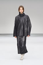 Placeholder for: Model wearing a black leather outfit with a high collar, walking on a runway against a plain white background.