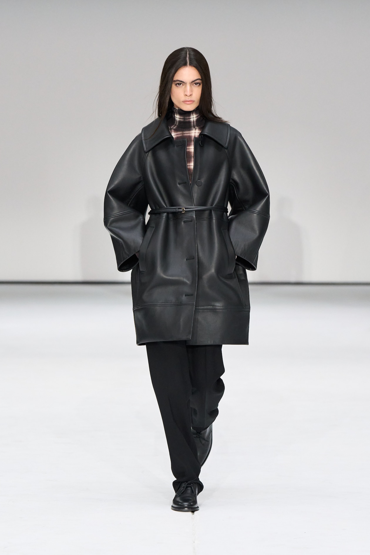 Model wearing a black leather coat over a plaid top and black pants, walking on a white runway.