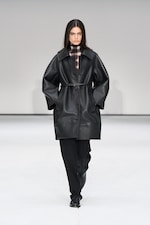 Placeholder for: Model wearing a black leather coat over a plaid top and black pants, walking on a white runway.