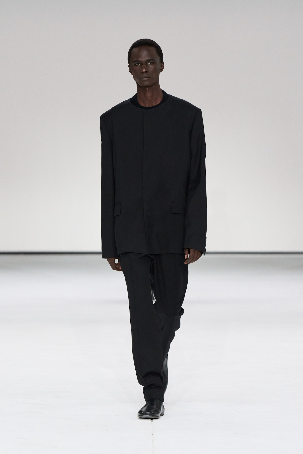 Person walking on a runway wearing an oversized black suit and black shoes against a plain white background.