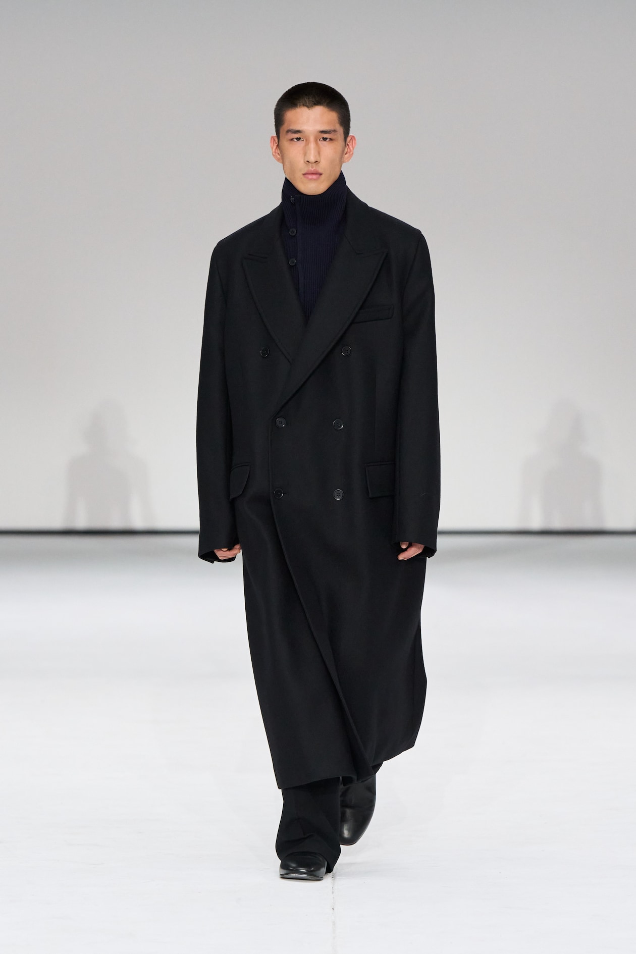 A person walks down a runway wearing a long black coat, black pants, and a dark turtleneck, set against a plain white background.