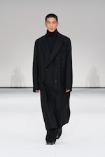 Placeholder for: A person walks down a runway wearing a long black coat, black pants, and a dark turtleneck, set against a plain white background.