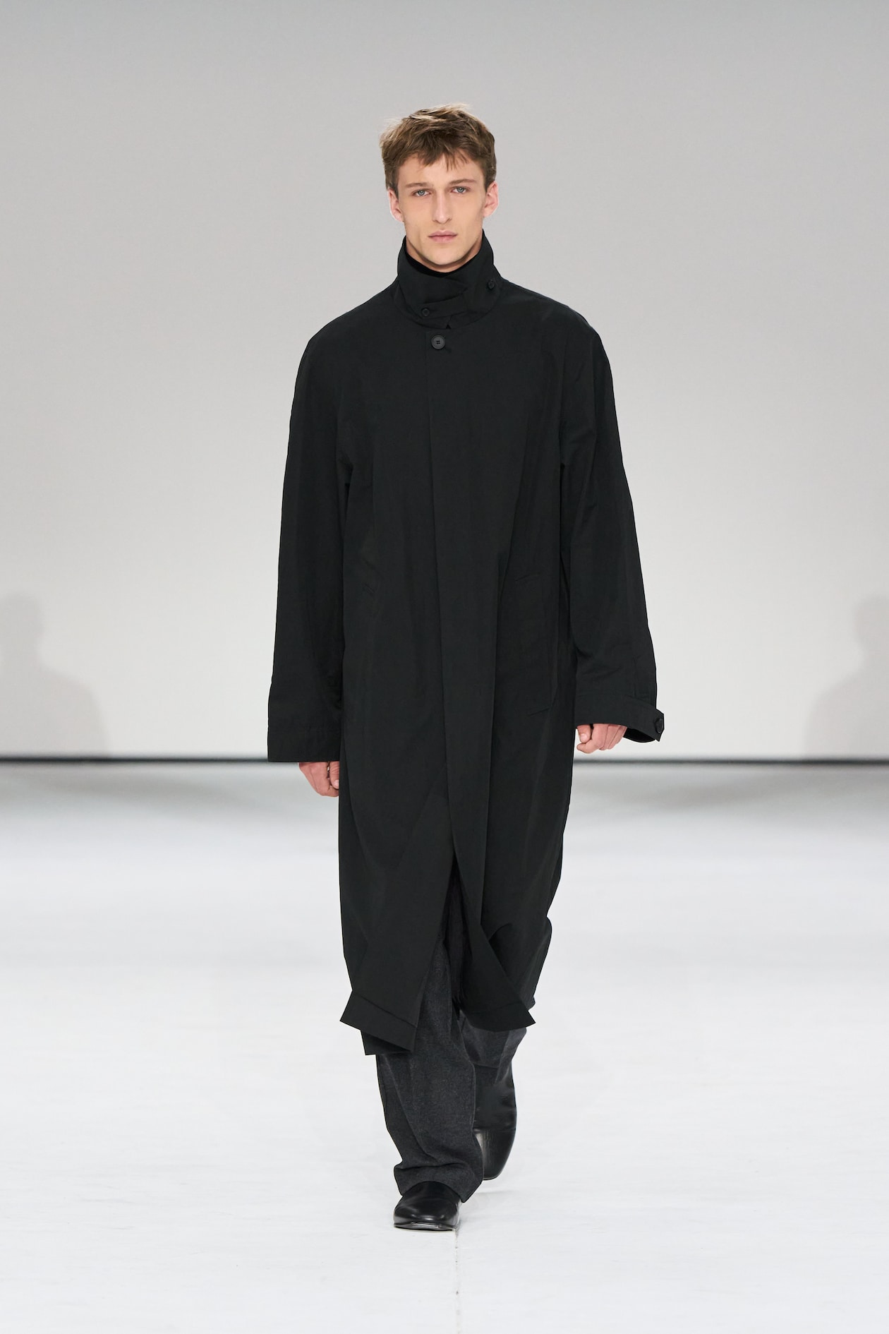 A person in a long black coat and dark pants walks confidently on a runway with a plain gray background.