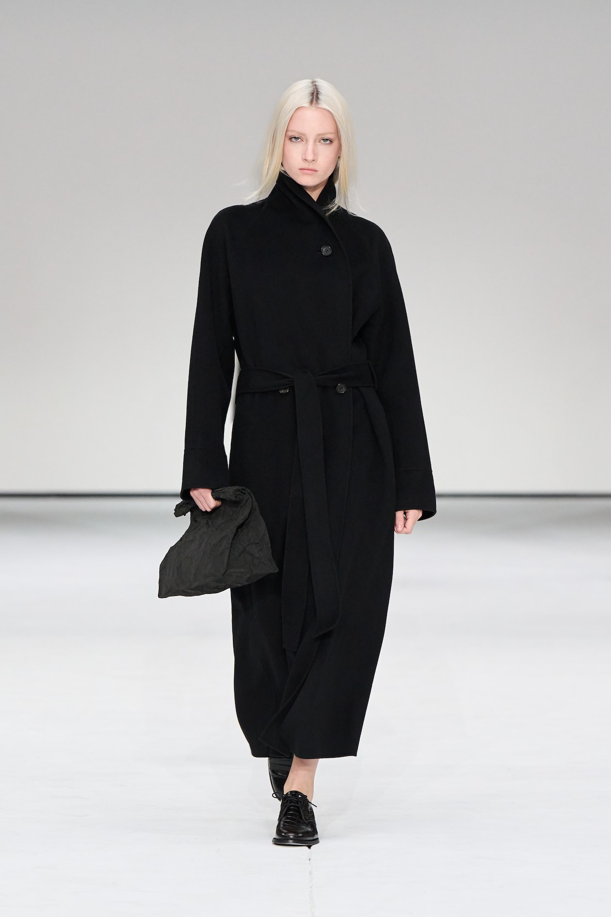 Model in a long black coat walks the runway, holding a black clutch, with a neutral expression and straight blonde hair.
