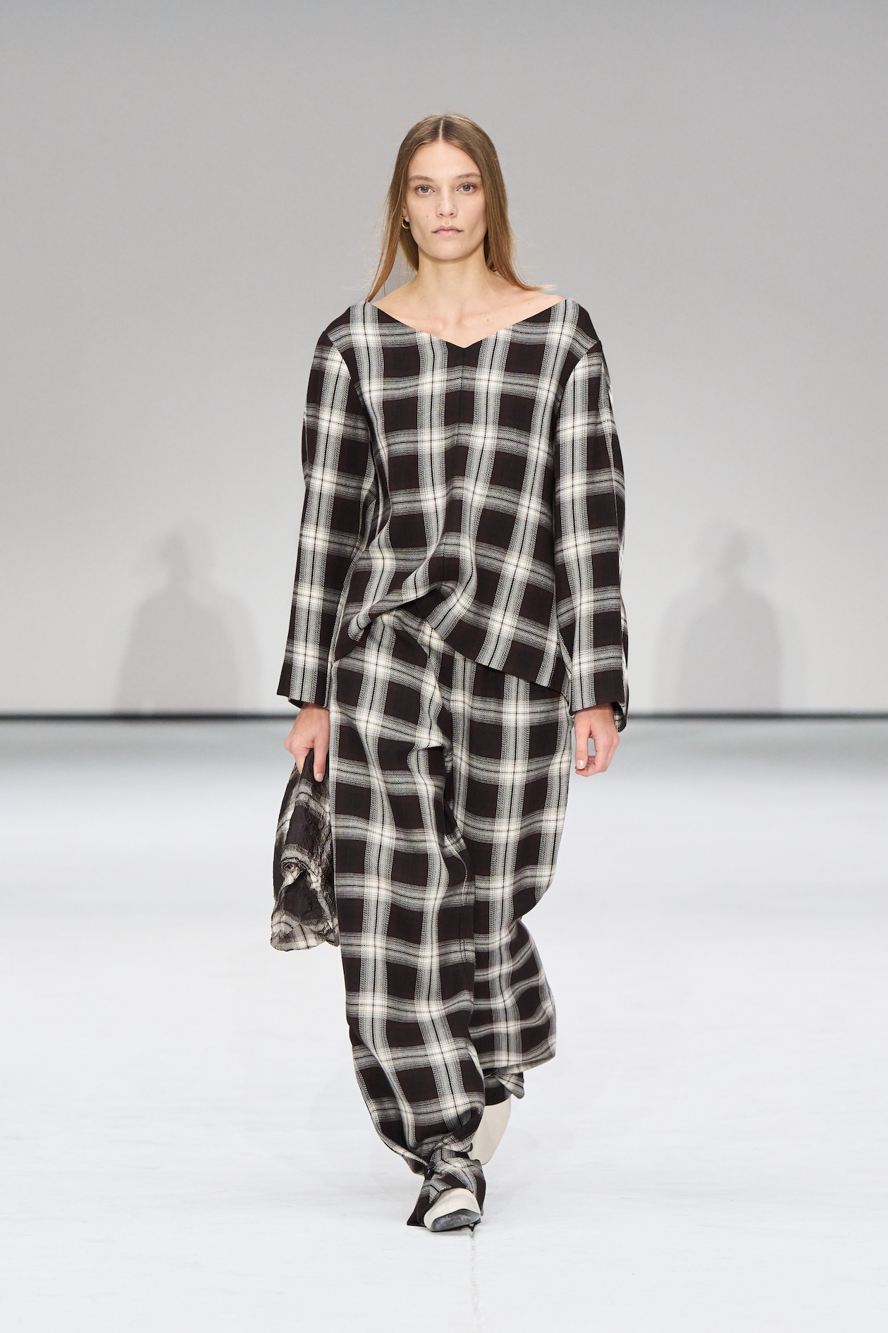 A model walks the runway wearing a loose-fitting black and white plaid outfit, holding a small bag, against a plain background.