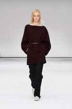 Placeholder for: Model in a burgundy oversized top, black pants, and white shoes walks on a minimalist runway.