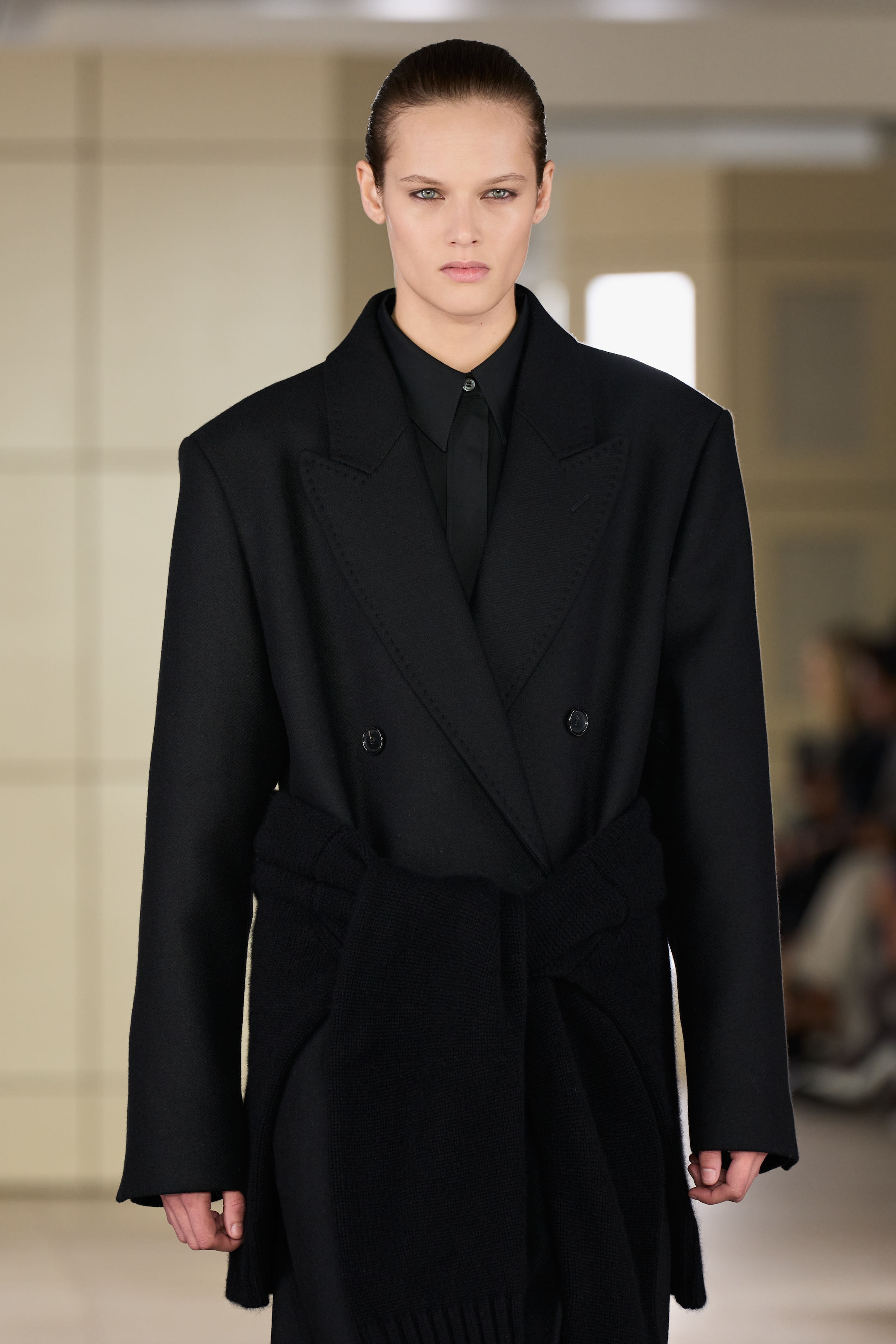 Person wearing a black oversized coat with a matching belt, walking down a runway. The background is neutral, with blurred spectators.