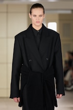 Placeholder for: Person wearing a black oversized coat with a matching belt, walking down a runway. The background is neutral, with blurred spectators.