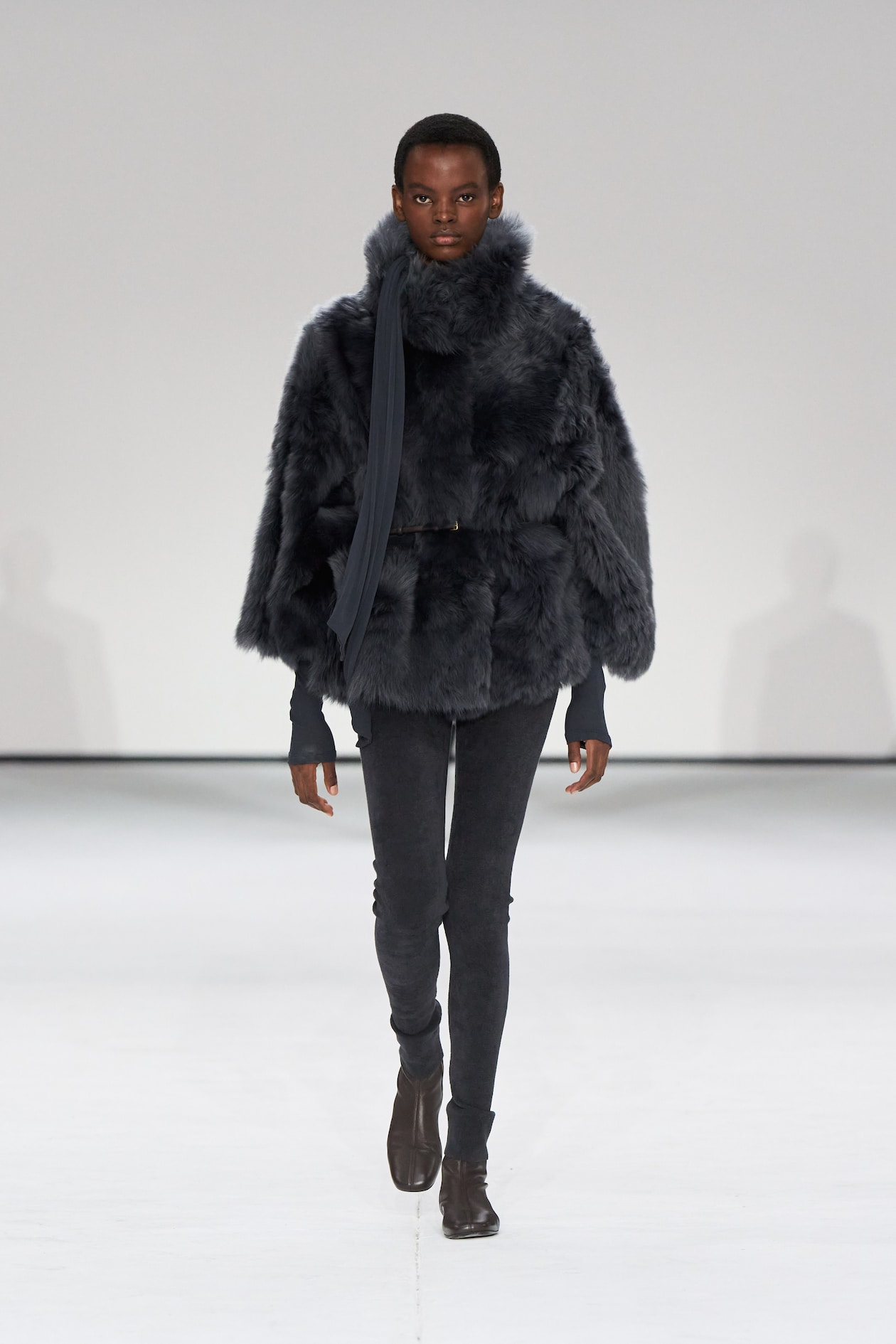 Model in a textured dark fur coat, scarf, and slim pants walks on a minimalist runway with a white background.