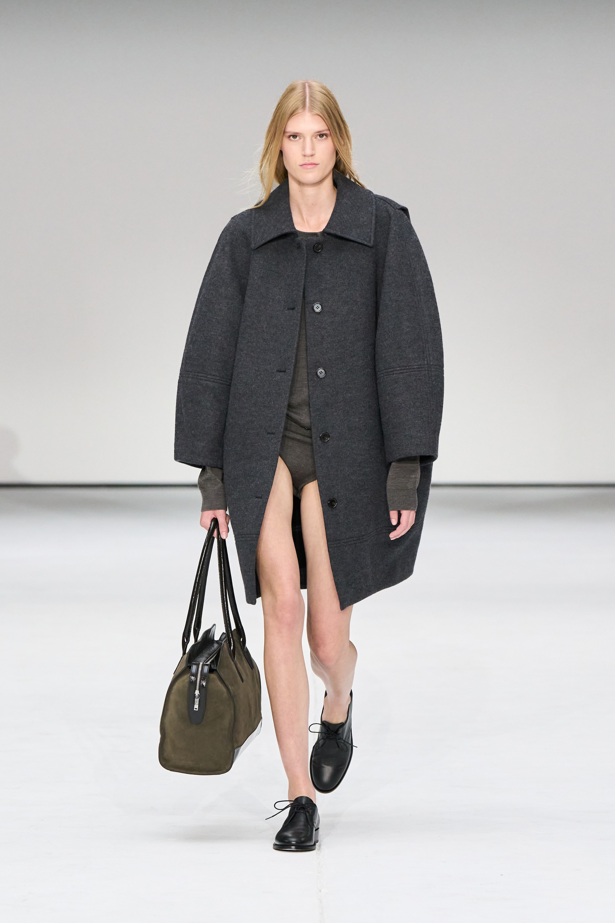 Model walking on runway wearing a dark oversized coat, black shoes, and carrying a large olive-green bag against a plain background.
