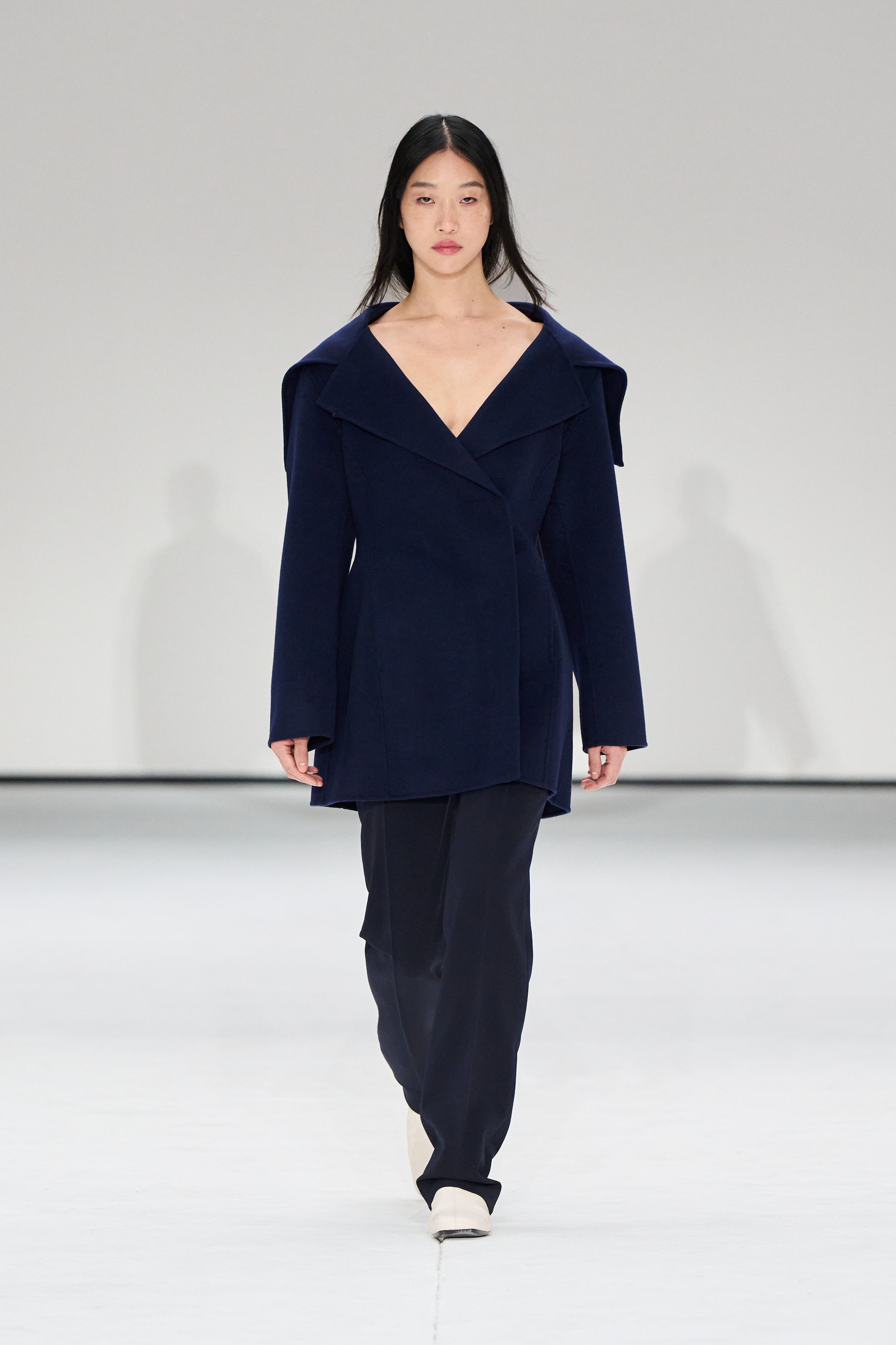 A model walks the runway wearing a navy blue coat with wide lapels and black pants, set against a plain white background.