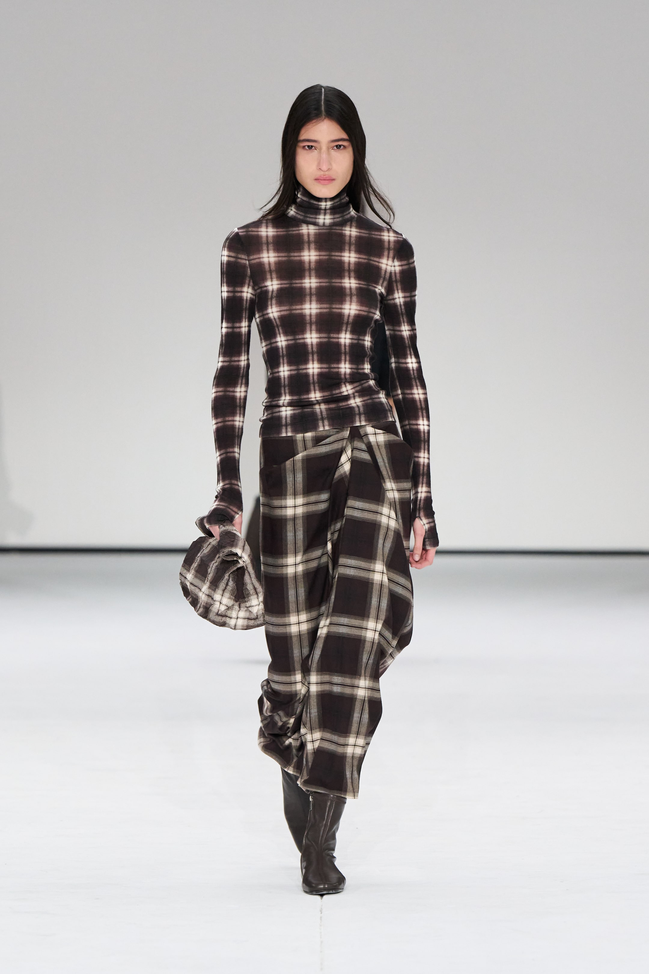 Model in plaid turtleneck and skirt ensemble walks runway, holding matching accessory. White background.