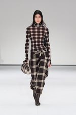 Placeholder for: Model in plaid turtleneck and skirt ensemble walks runway, holding matching accessory. White background.