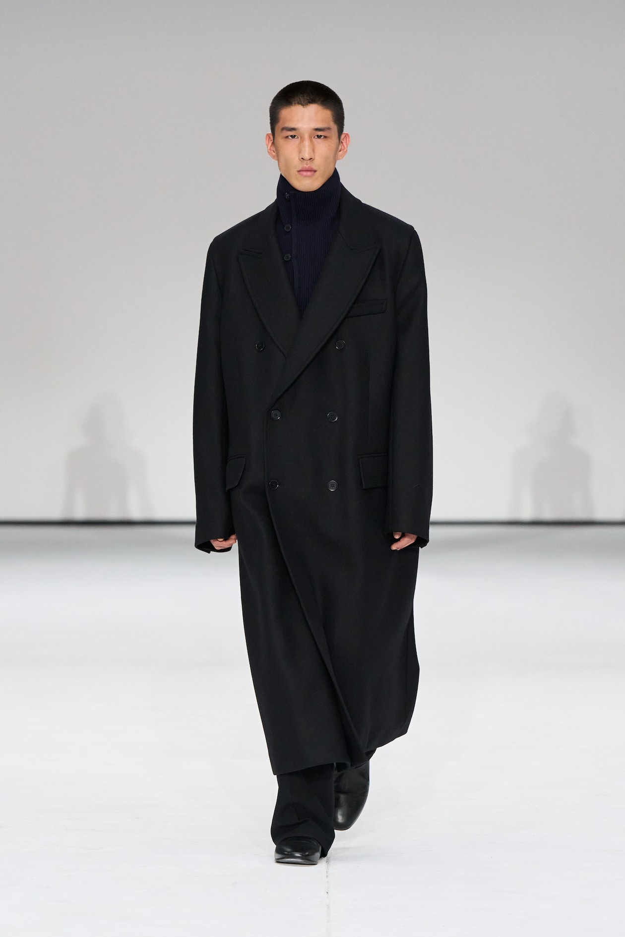 Model wearing a long black coat over a dark turtleneck and pants, walking on a white runway.