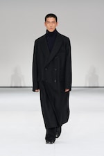 Placeholder for: Model wearing a long black coat over a dark turtleneck and pants, walking on a white runway.
