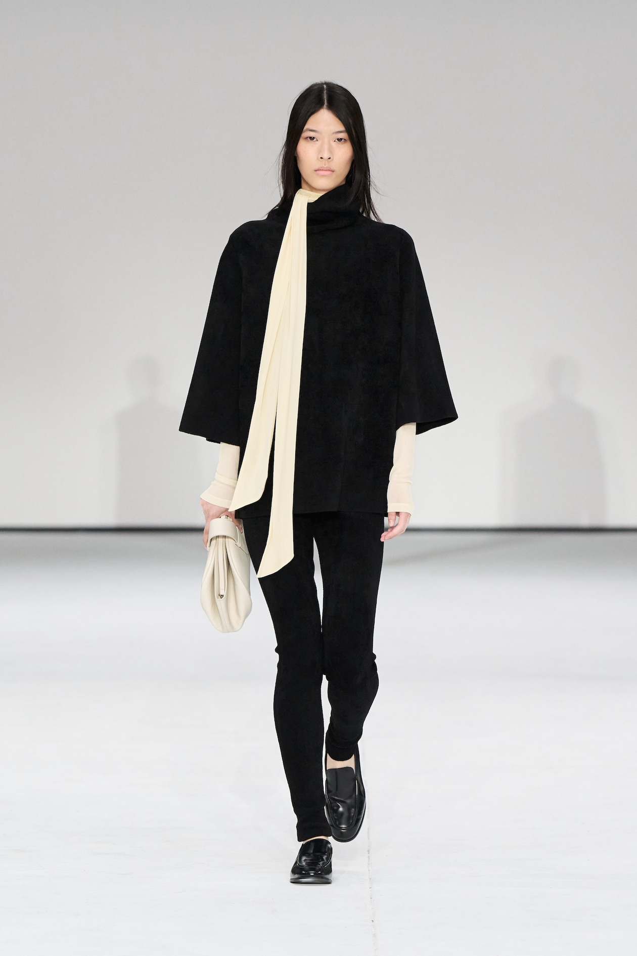 Model in black outfit with a cream scarf walks on a runway, holding a matching cream clutch. Minimalist fashion show setting.
