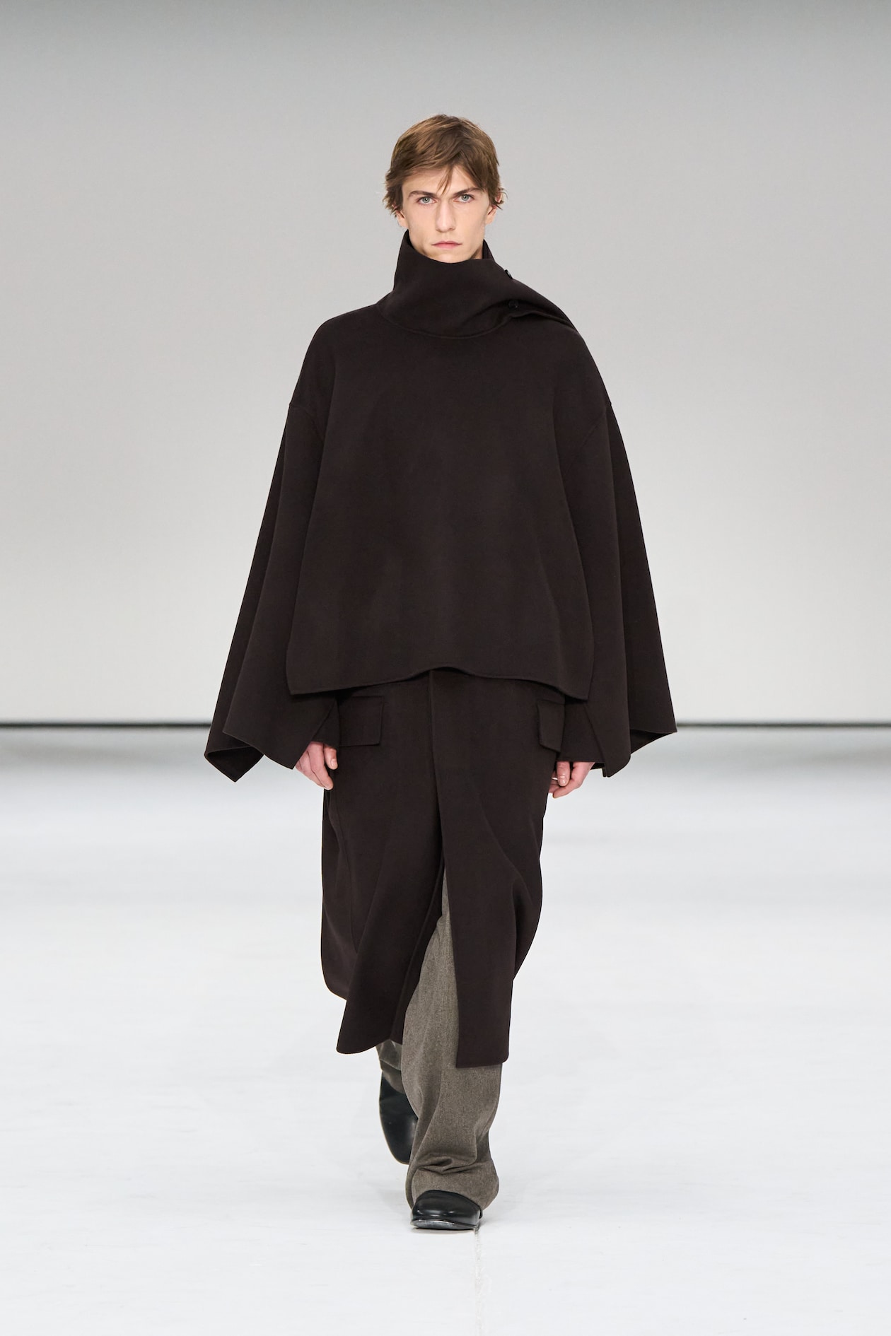 Model wearing a dark, layered poncho-style outfit and boots, walking on a white runway with a neutral background.