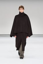 Placeholder for: Model wearing a dark, layered poncho-style outfit and boots, walking on a white runway with a neutral background.