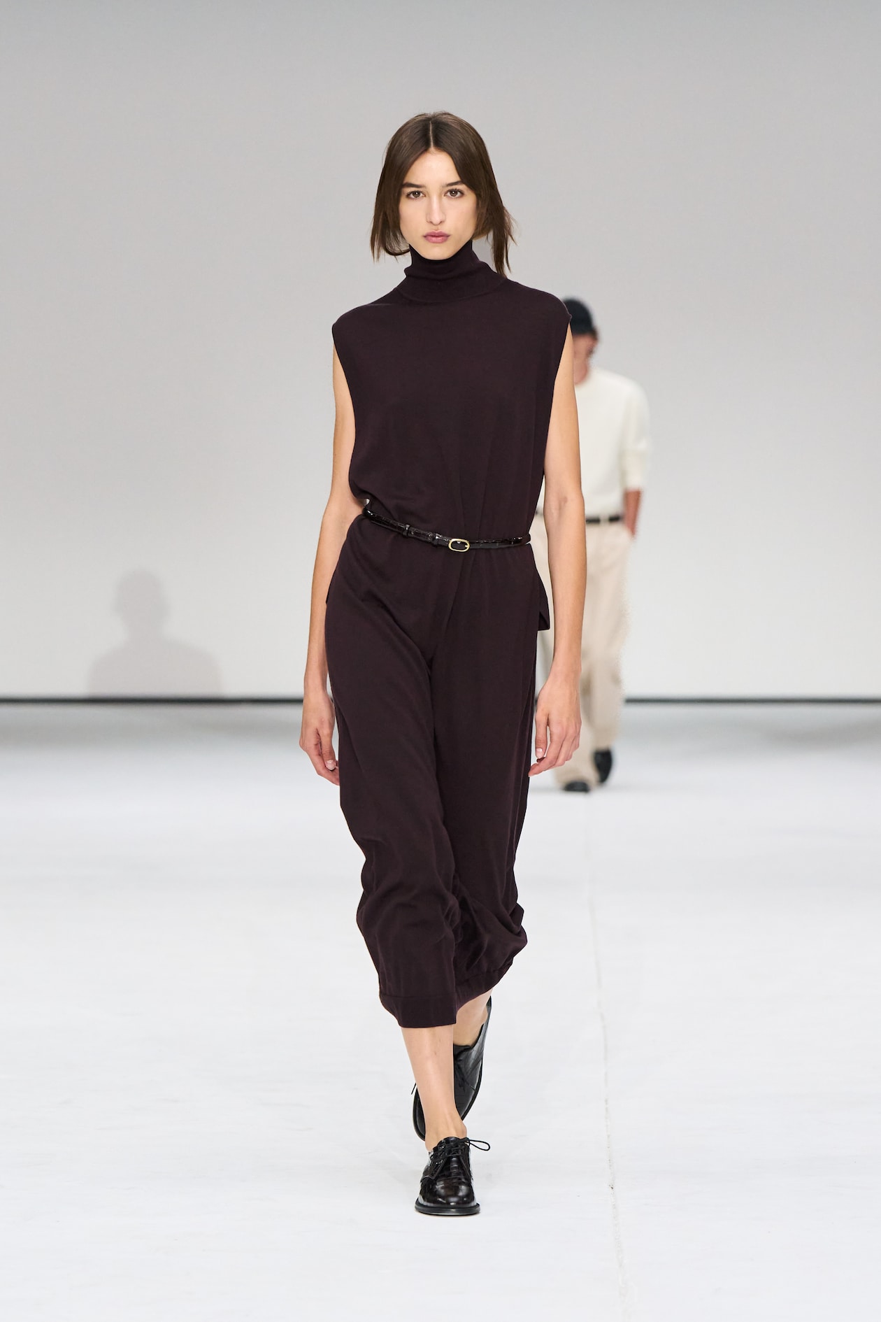 Model in a sleeveless black jumpsuit and black shoes walks on a white runway, with another person in the background.
