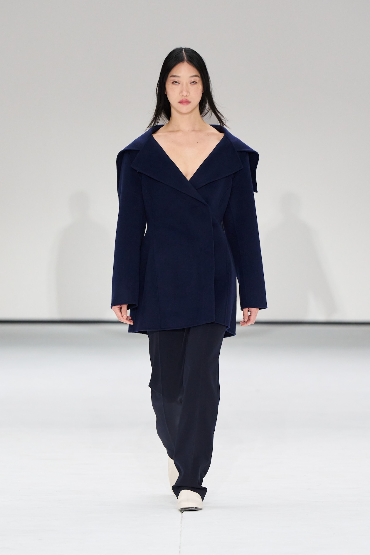 Model in a navy blue outfit with a wide-collared coat and pants walks on a white runway.