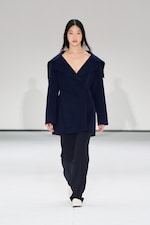Placeholder for: Model in a navy blue outfit with a wide-collared coat and pants walks on a white runway.