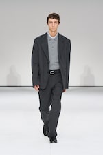Placeholder for: Man in a gray suit and polo shirt walks confidently on a white runway, showcasing a fashion design.