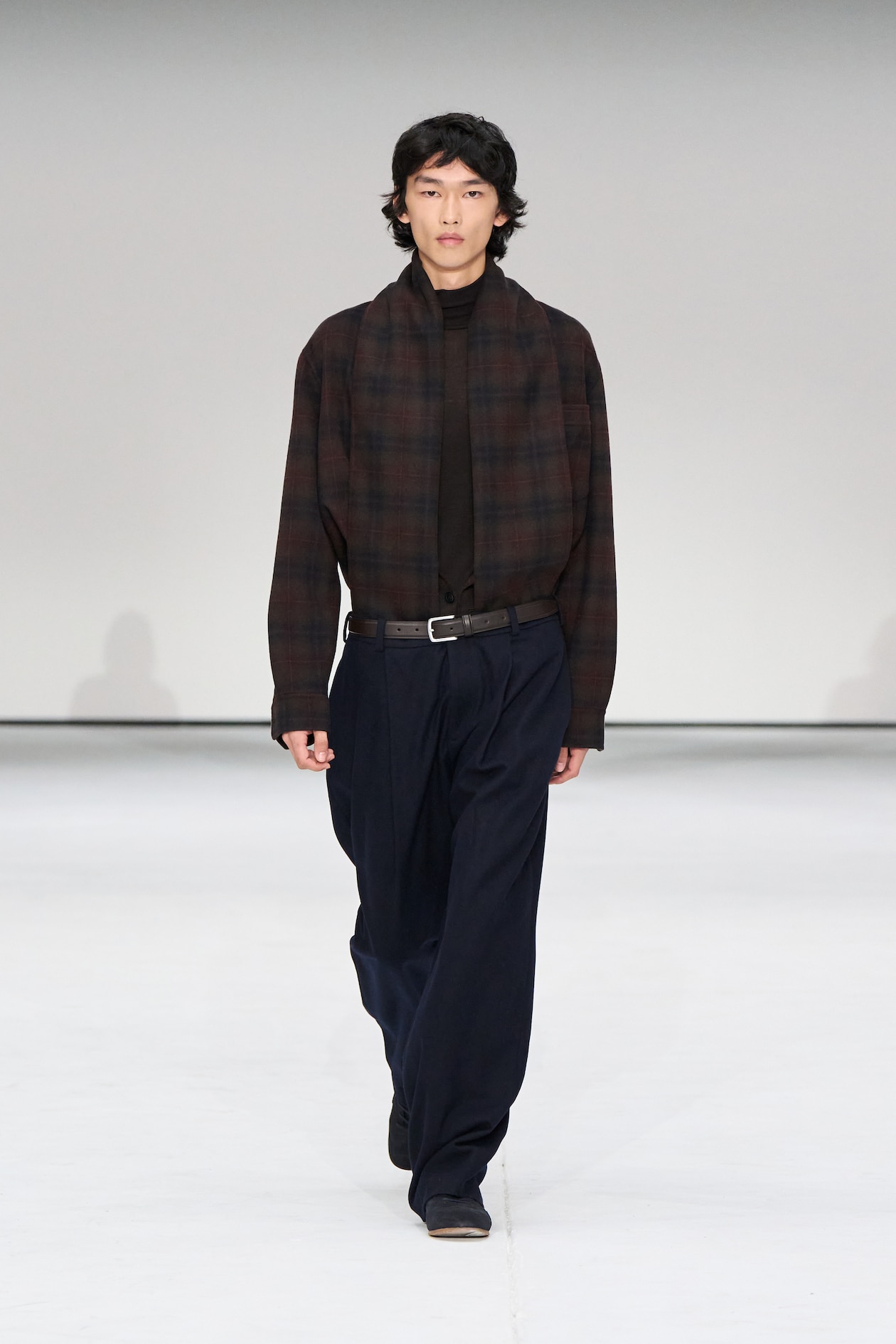 Model walking on runway wearing a dark plaid shirt, black scarf, and loose navy pants against a plain white background.