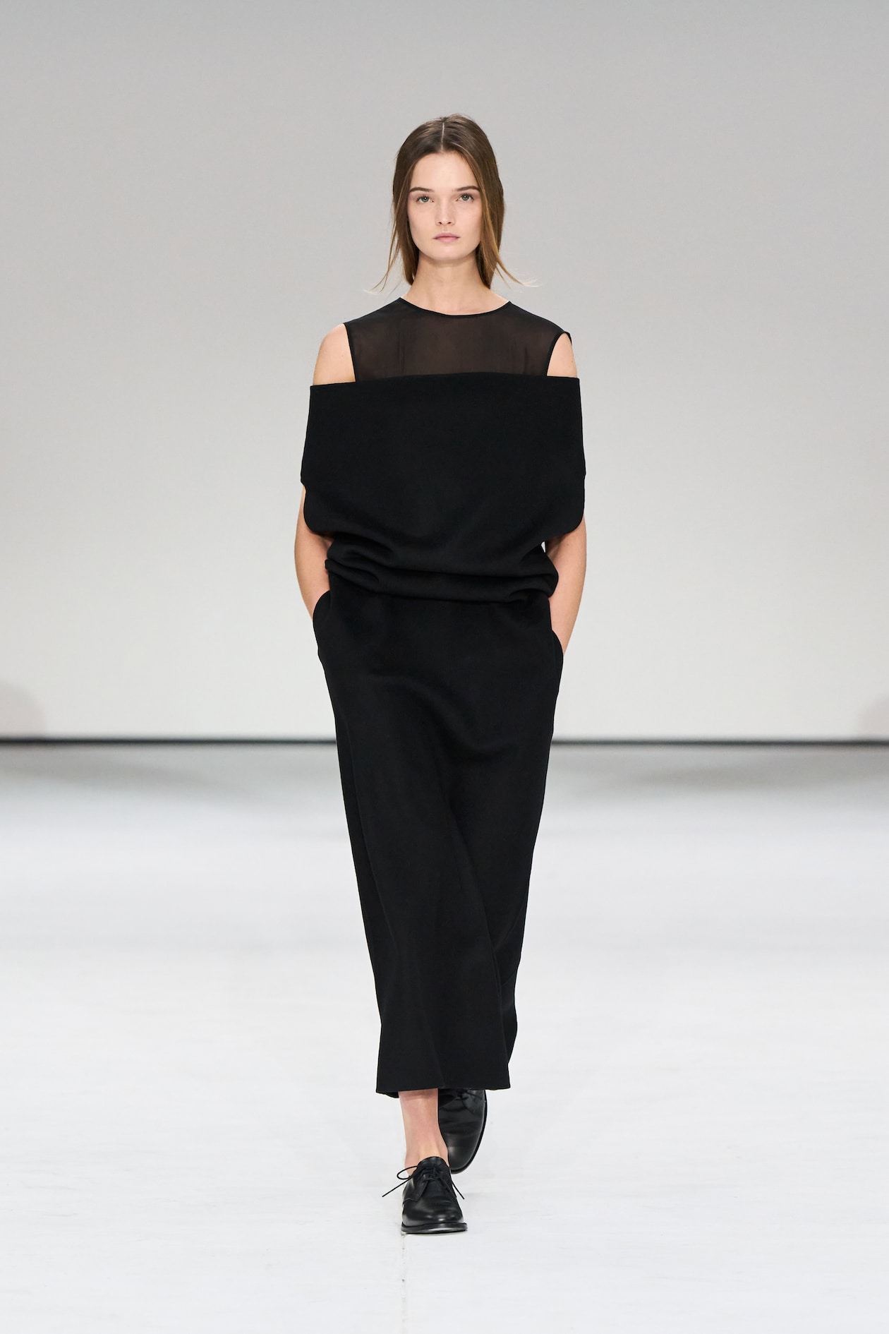 Model in a black, off-shoulder dress with mesh top, walking on a white runway, hands in pockets, wearing black shoes.