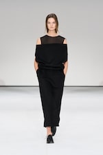 Placeholder for: Model in a black, off-shoulder dress with mesh top, walking on a white runway, hands in pockets, wearing black shoes.