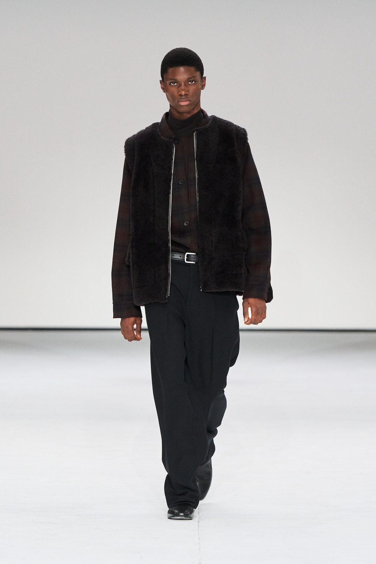 Model in a dark fur vest, plaid shirt, and black pants walks on a fashion runway against a plain white background.