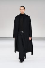 Placeholder for: Model in a long black coat and black outfit walks down a minimalist runway with a neutral background.