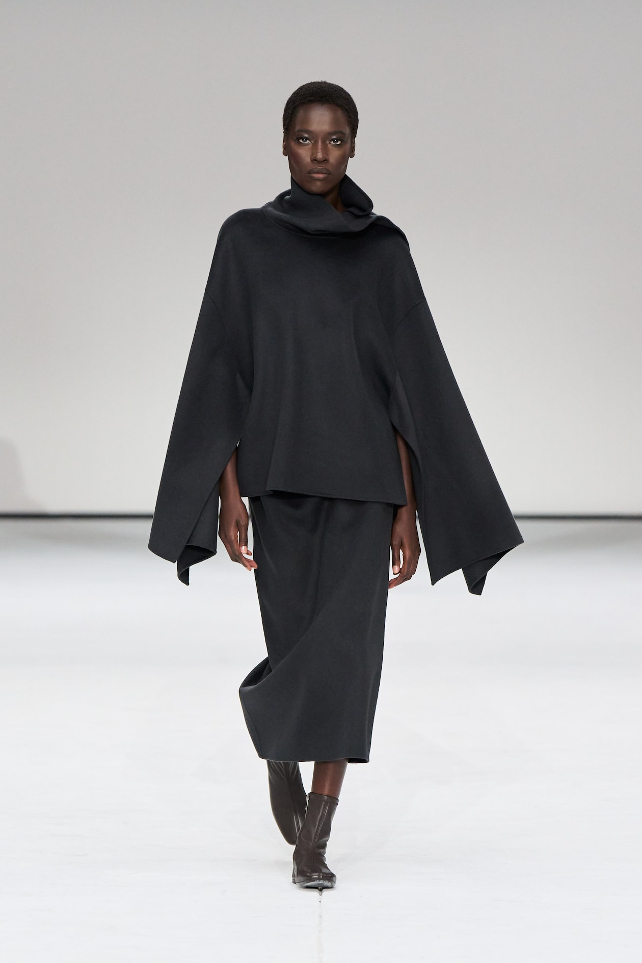 Model wearing a dark, cape-style outfit with a high collar and knee-length skirt, striding confidently on a minimalist runway.