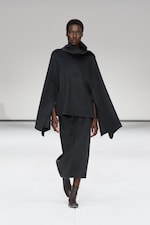 Placeholder for: Model wearing a dark, cape-style outfit with a high collar and knee-length skirt, striding confidently on a minimalist runway.