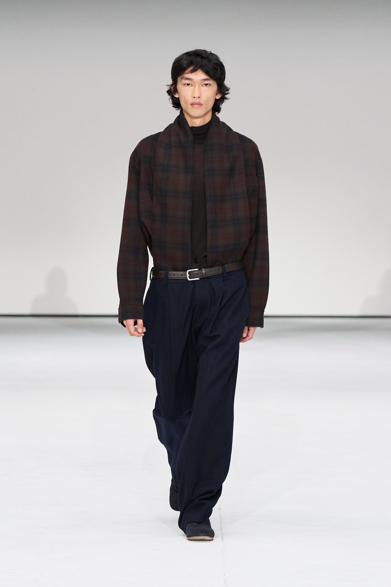 Model in a dark plaid shirt and loose navy pants walks on a white runway.