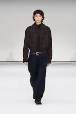 Placeholder for: Model in a dark plaid shirt and loose navy pants walks on a white runway.