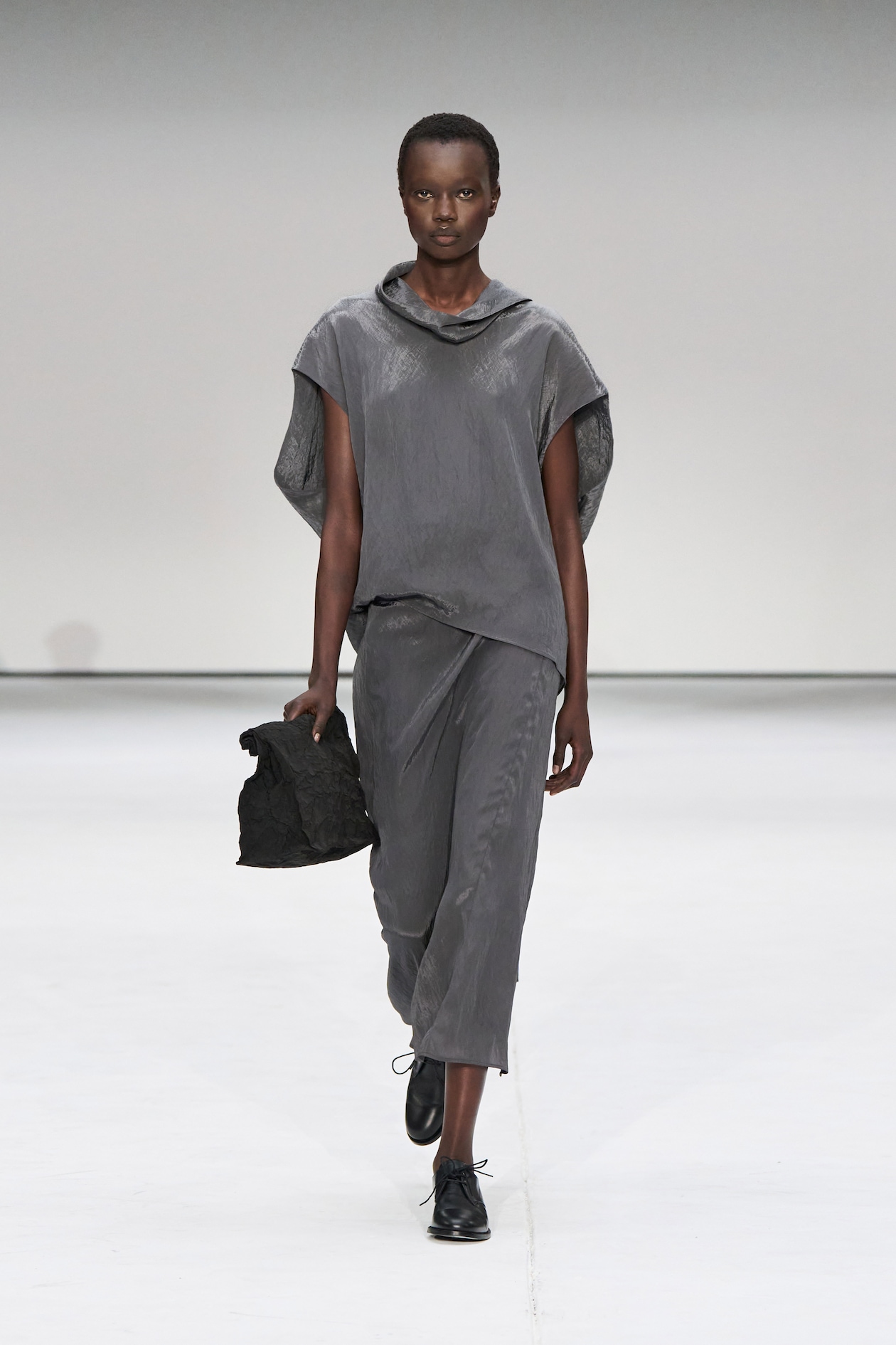 Model in a gray, asymmetrical outfit with matching pants, holding a black bag, walks on a white runway.