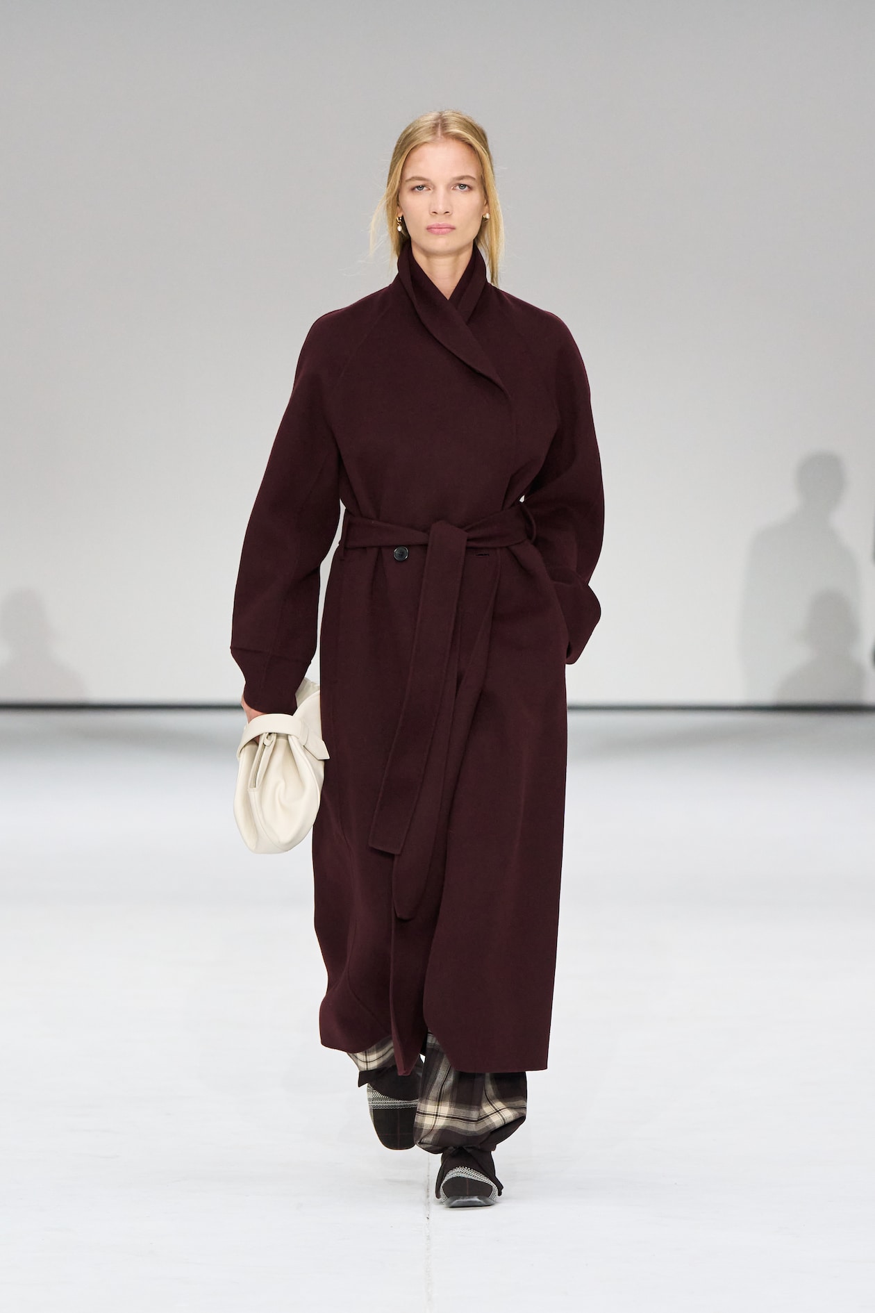 A model walks the runway in a long, dark brown coat with a belt, holding a white clutch, against a plain background.