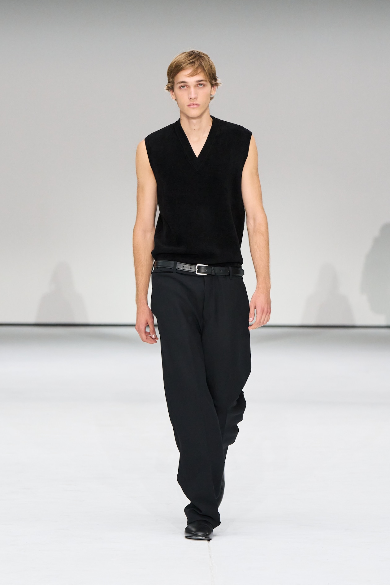 A model in a sleeveless black top and black pants walks down a white runway during a fashion show.