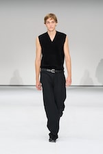 Placeholder for: A model in a sleeveless black top and black pants walks down a white runway during a fashion show.
