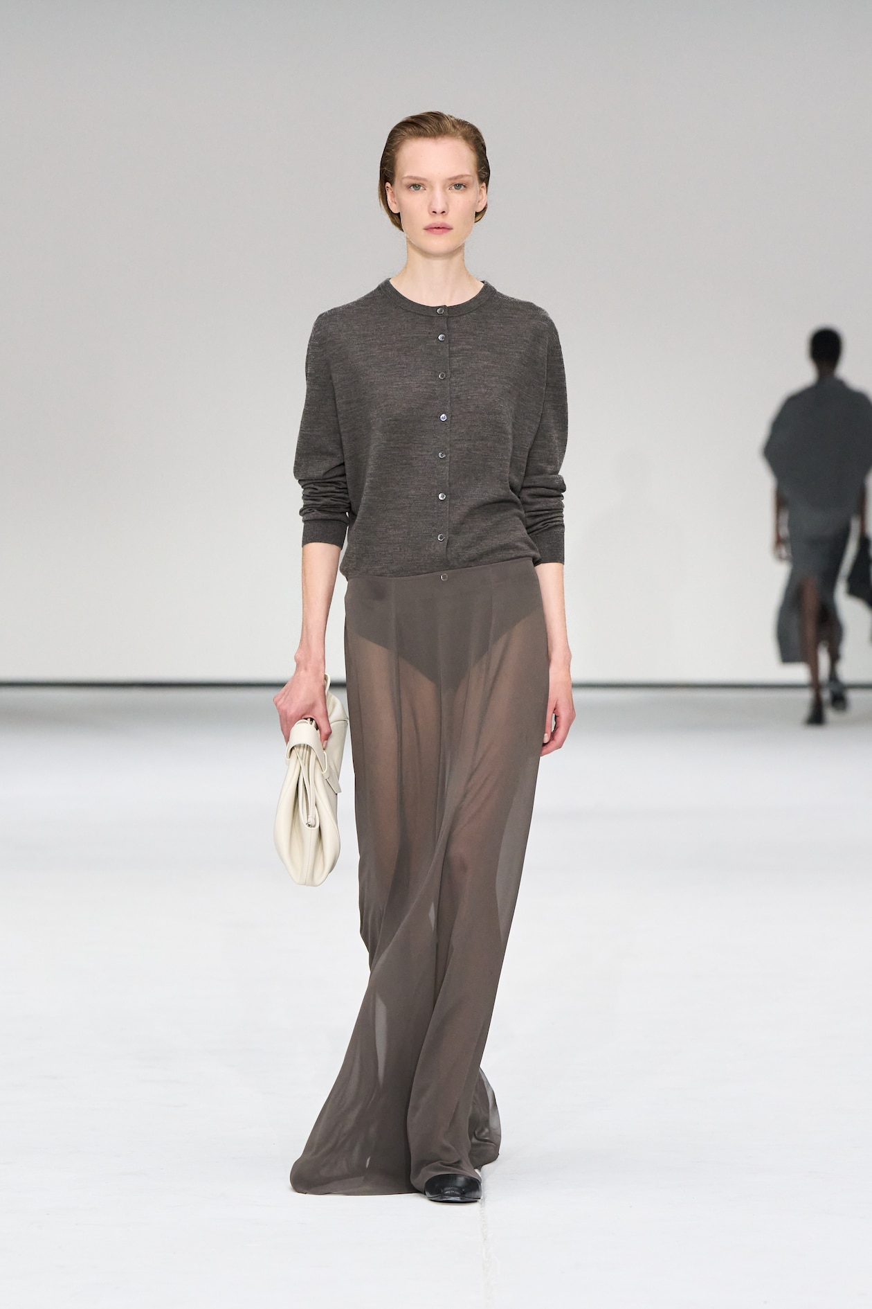 Model in a gray cardigan and sheer skirt walks the runway, holding a white clutch, against a minimalist white backdrop.