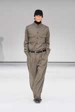 Placeholder for: Model in a gray button-up shirt and wide-leg trousers, wearing a black cap, walking on a runway with a neutral background.