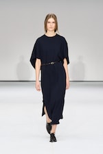 Placeholder for: A person walks on a runway wearing a dark blue dress with a black belt and black shoes, set against a plain, light-colored background.